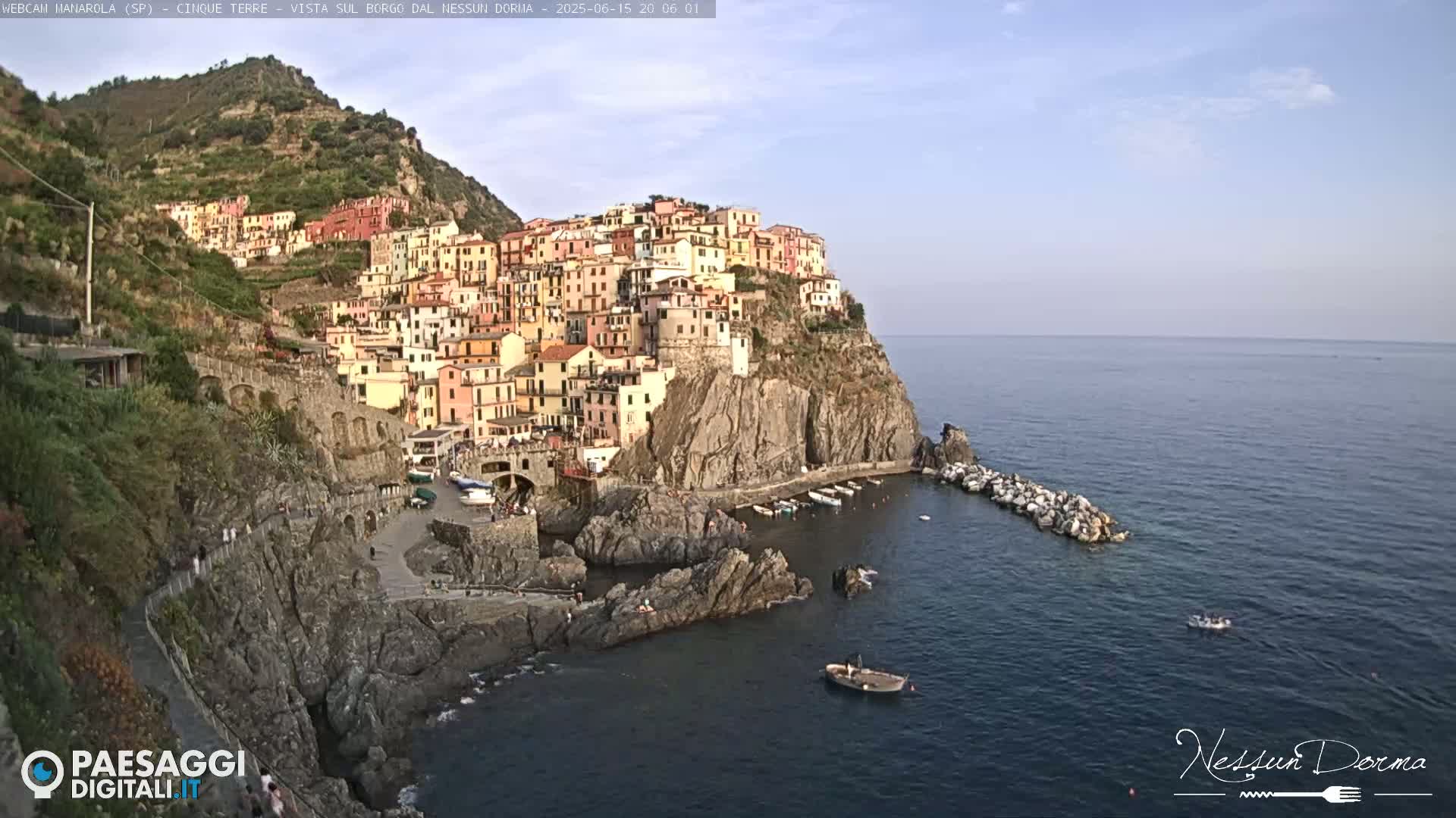 A colorful village clings to a steep cliffside overlooking a calm, dark-blue sea under a clear sky.