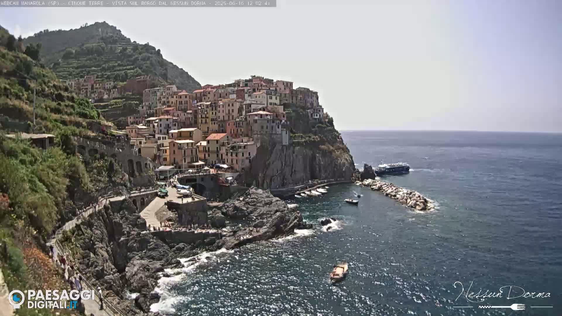 A colorful village clings to a steep cliffside overlooking a calm sea on a sunny day.