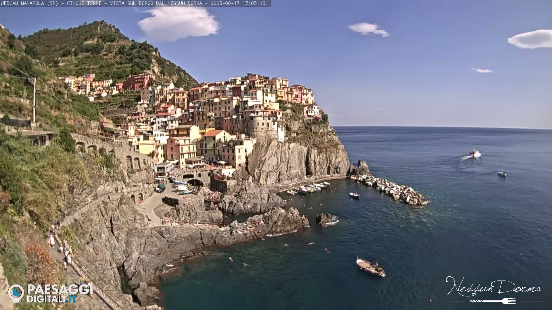 A colorful village clings to a steep cliffside overlooking a calm, blue sea with several small boats.
