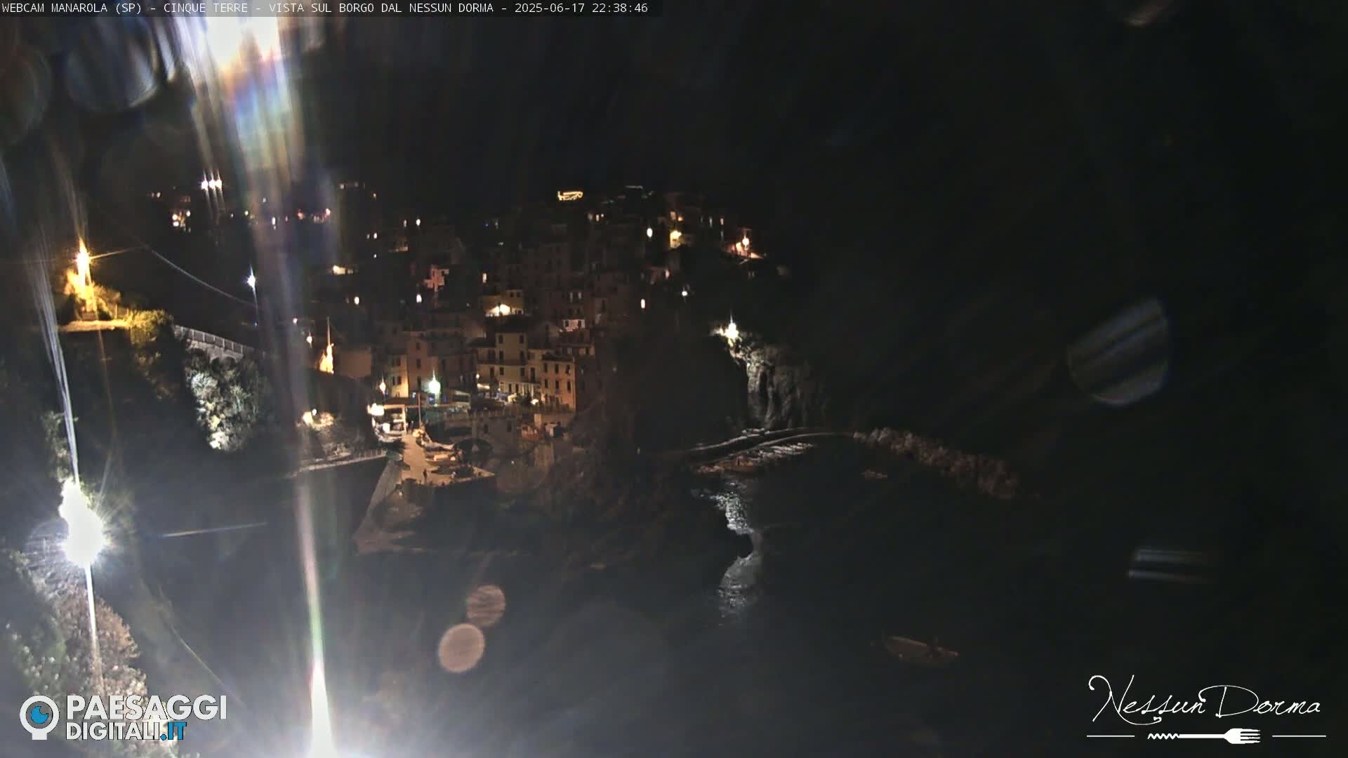 A nighttime, high-angle view of a coastal town nestled on a cliffside, illuminated by scattered lights, with dark water visible below.