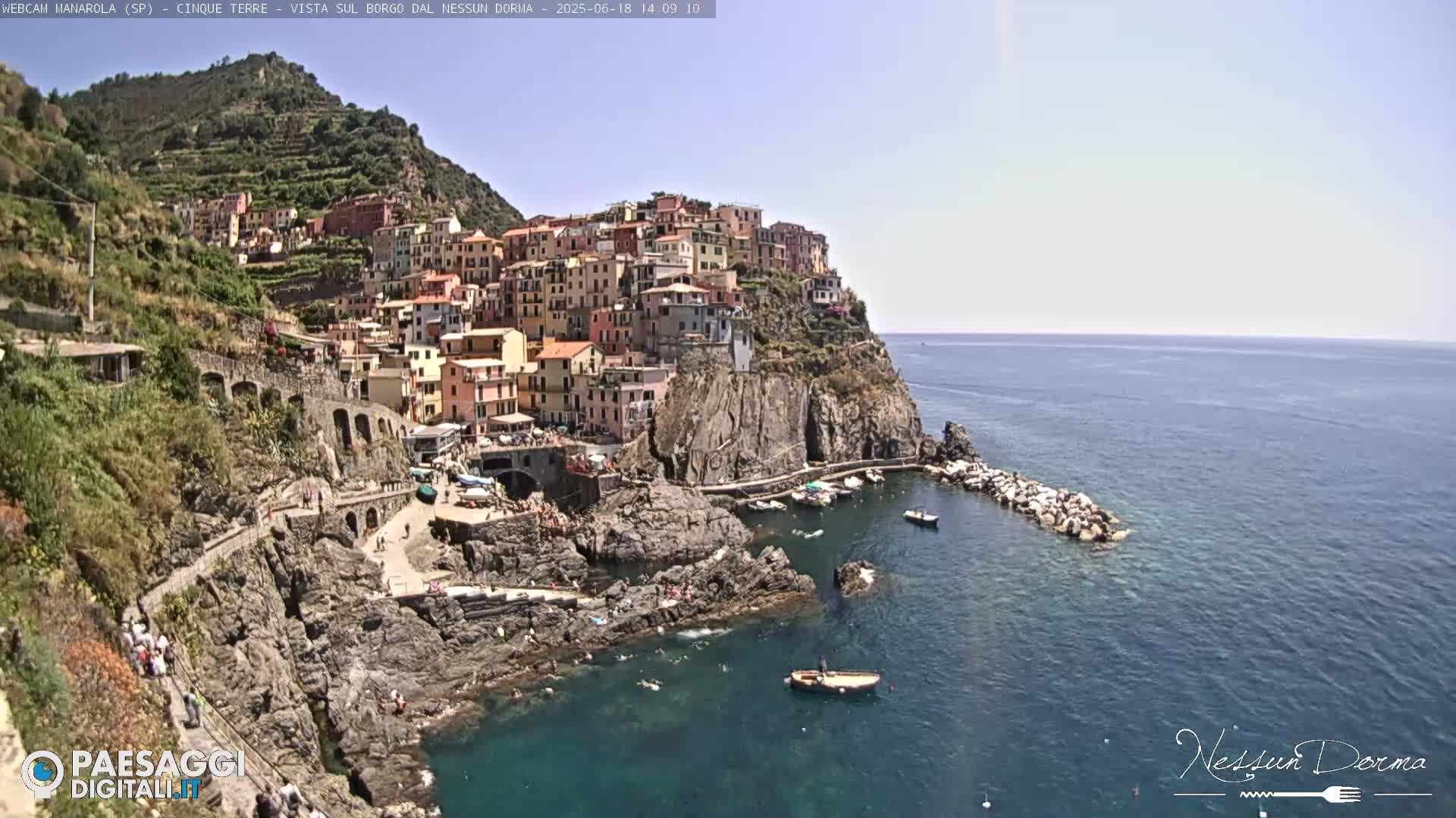 A colorful village clings to a steep cliffside overlooking a calm, blue sea on a sunny day.