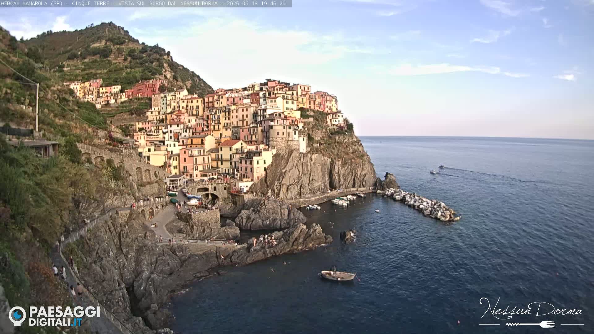 A colorful village clings to a steep cliffside overlooking a calm sea under a mostly clear sky.