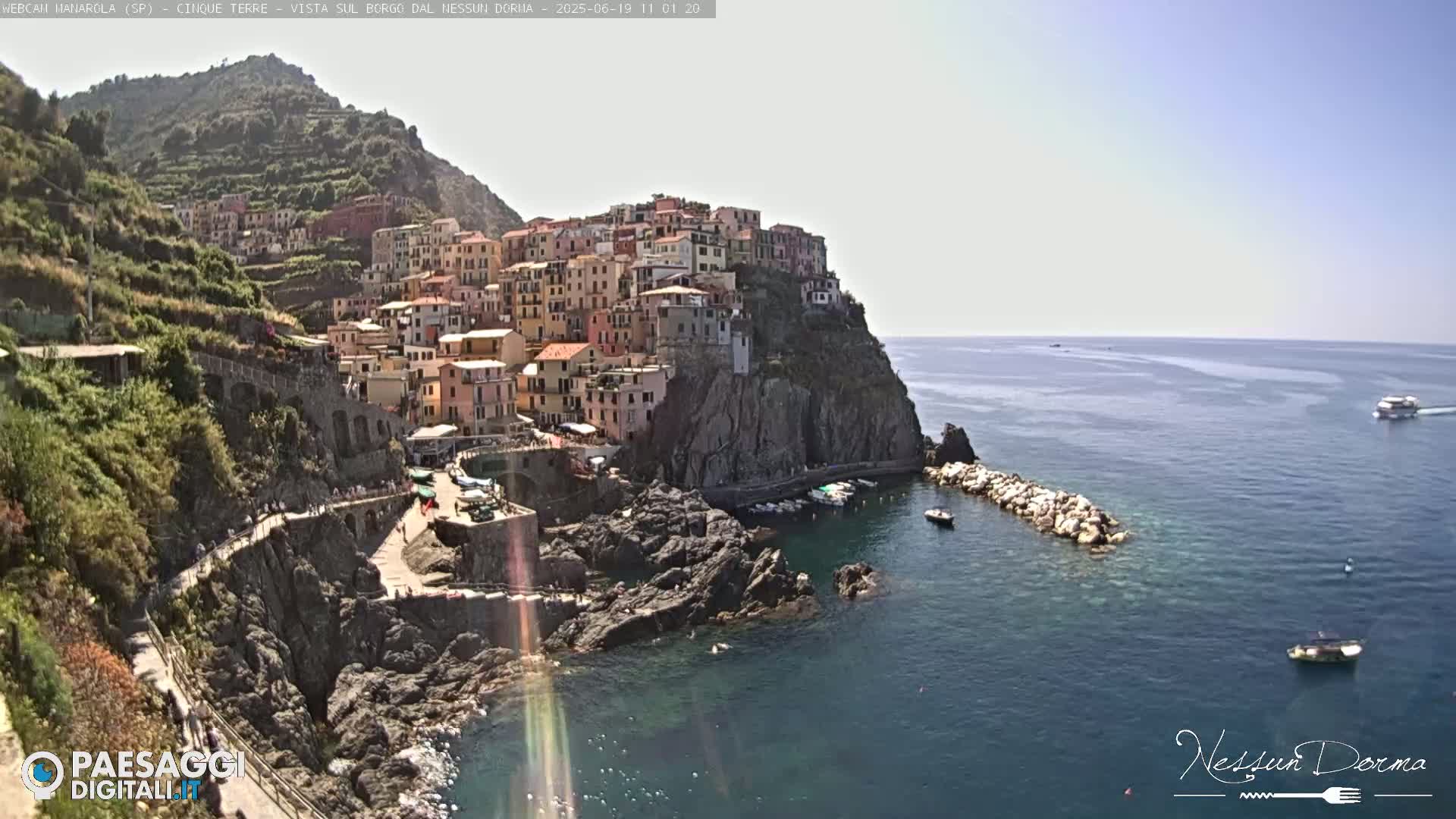 A colorful village clings to a rocky cliffside overlooking a calm, blue sea on a sunny day.