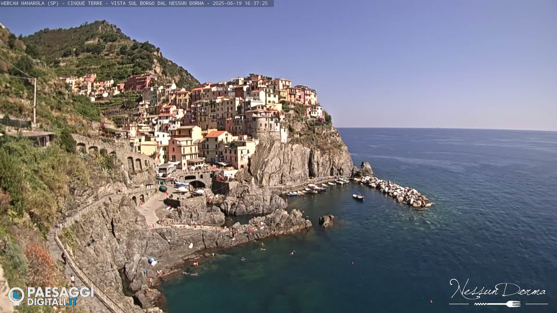 A colorful village clings to a rocky cliffside overlooking a calm, blue sea on a sunny day.