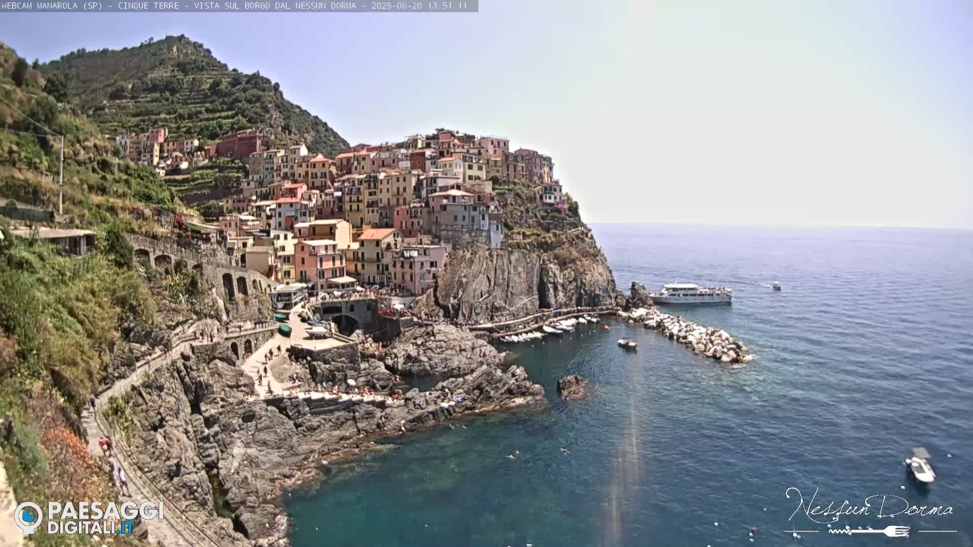 A colorful village clings to a steep cliffside overlooking a calm, clear sea where several boats are moored.