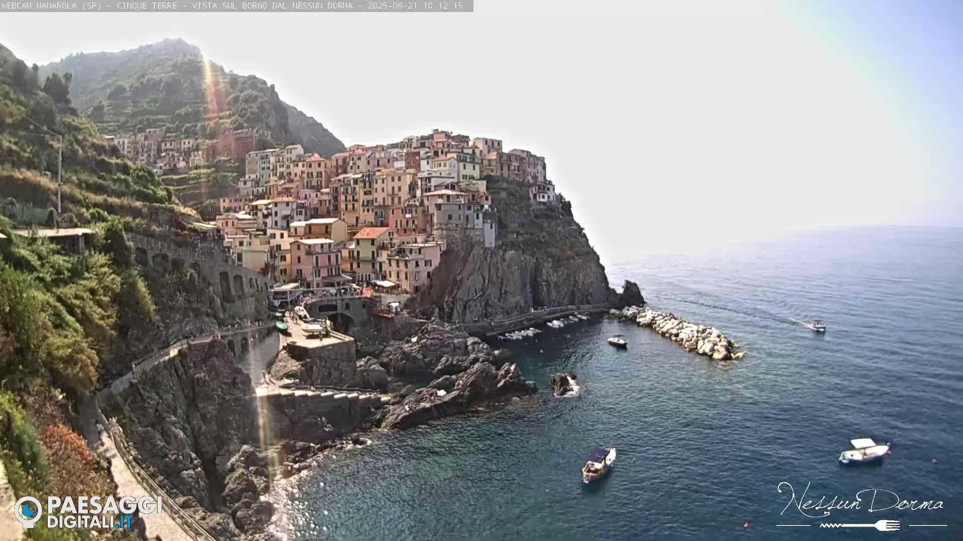 A colorful village clings to a rocky cliffside overlooking a calm blue sea, with a few small boats visible in the water.