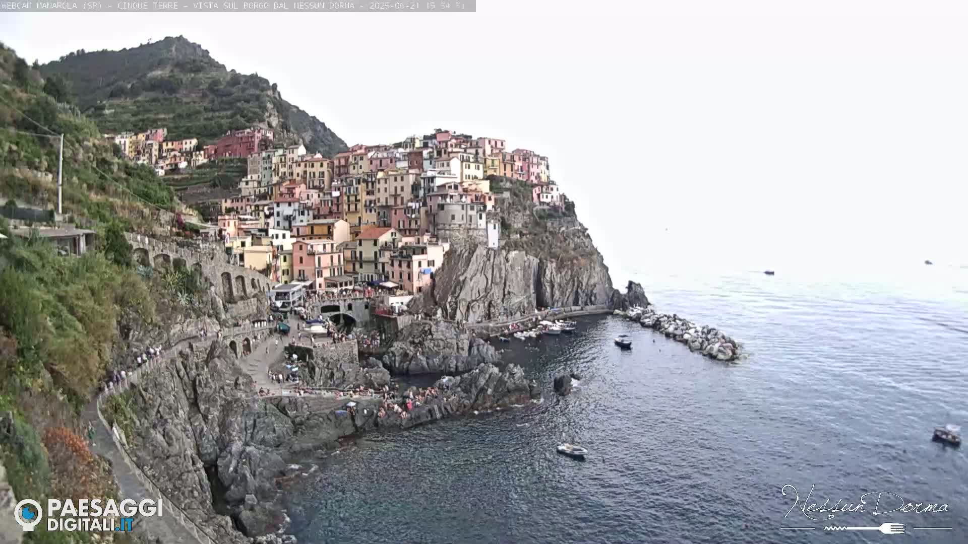 A colorful village clings to a rocky cliffside overlooking a calm sea with several small boats.