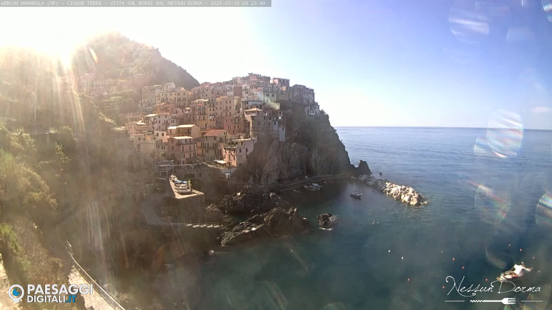 A brightly lit coastal town, nestled on a rocky cliffside, overlooks a calm sea under a clear sky.
