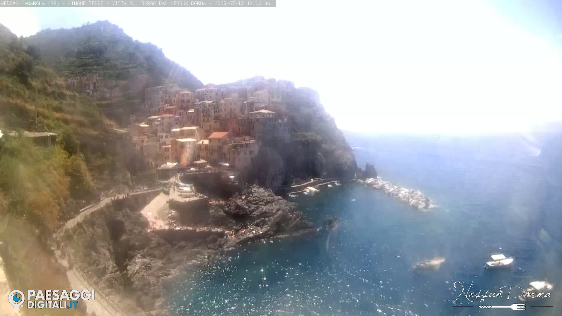 A brightly lit coastal town, nestled on a rocky cliffside, overlooks a calm sea under a clear sky.
