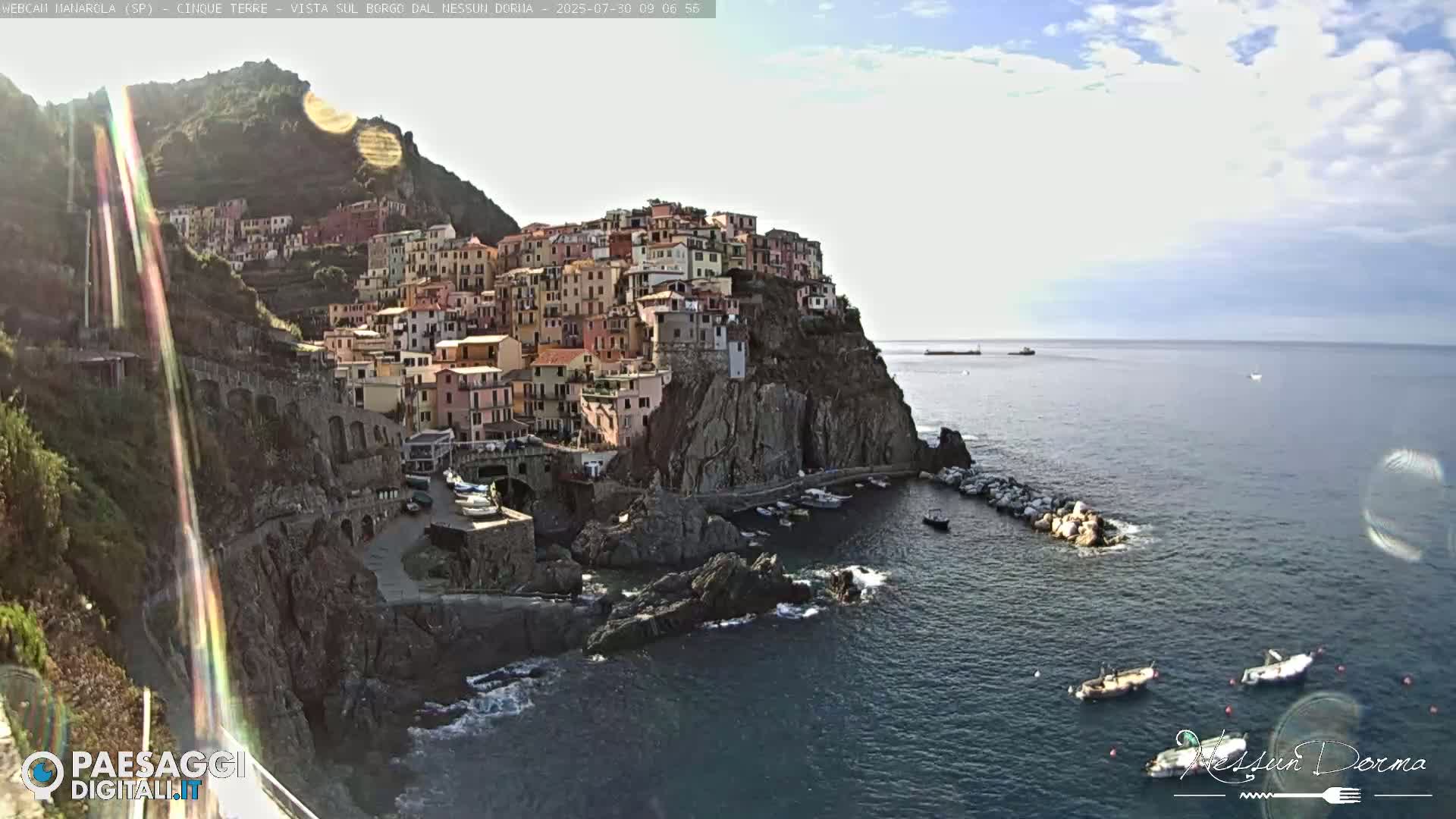 A colorful village clings to a steep cliffside overlooking a calm sea with several small boats.