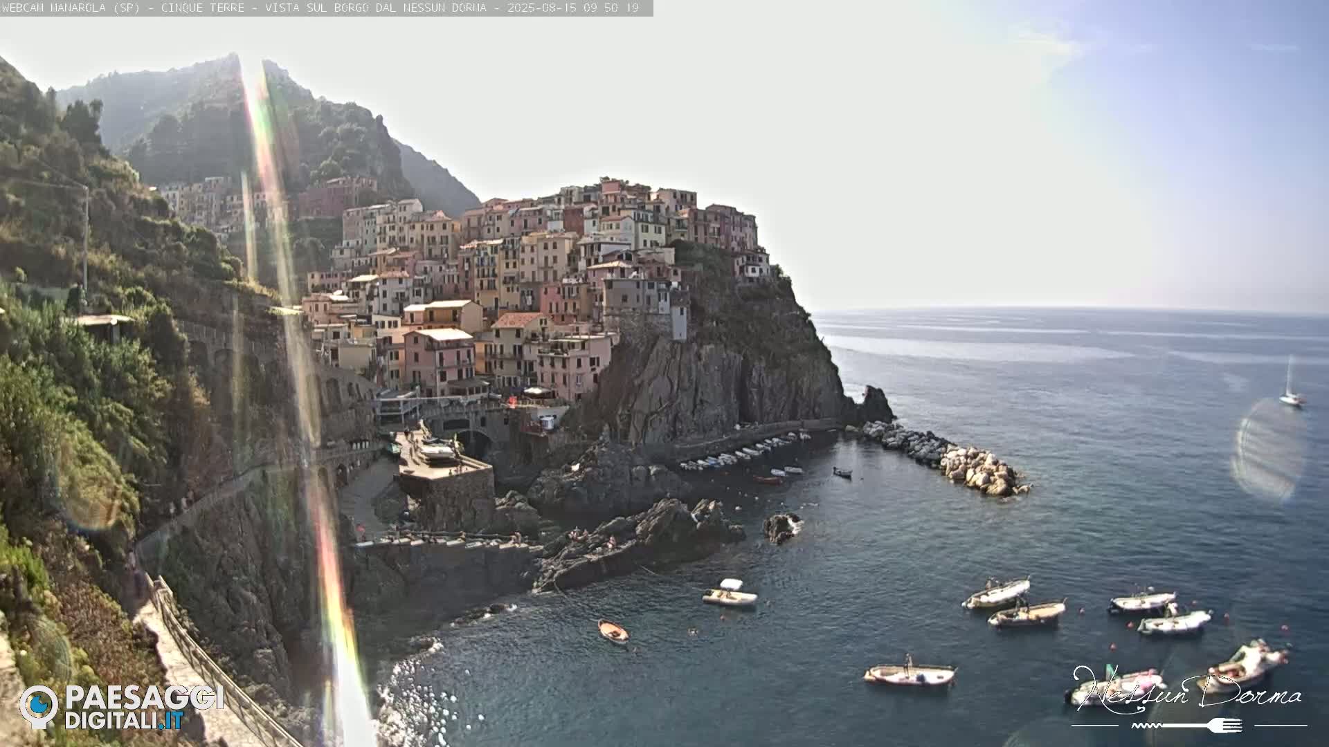 A picturesque coastal village clings to a steep cliffside overlooking a calm sea where several small boats are moored.