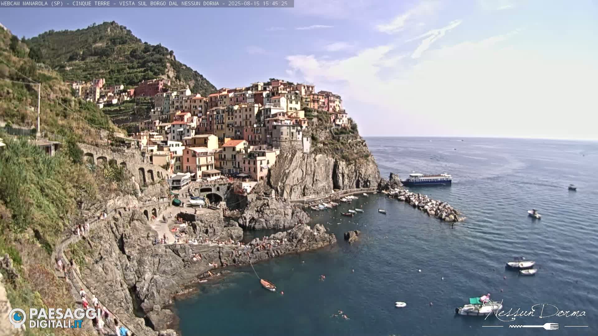 A picturesque coastal village clings to a steep cliffside overlooking a calm sea where several small boats are moored.