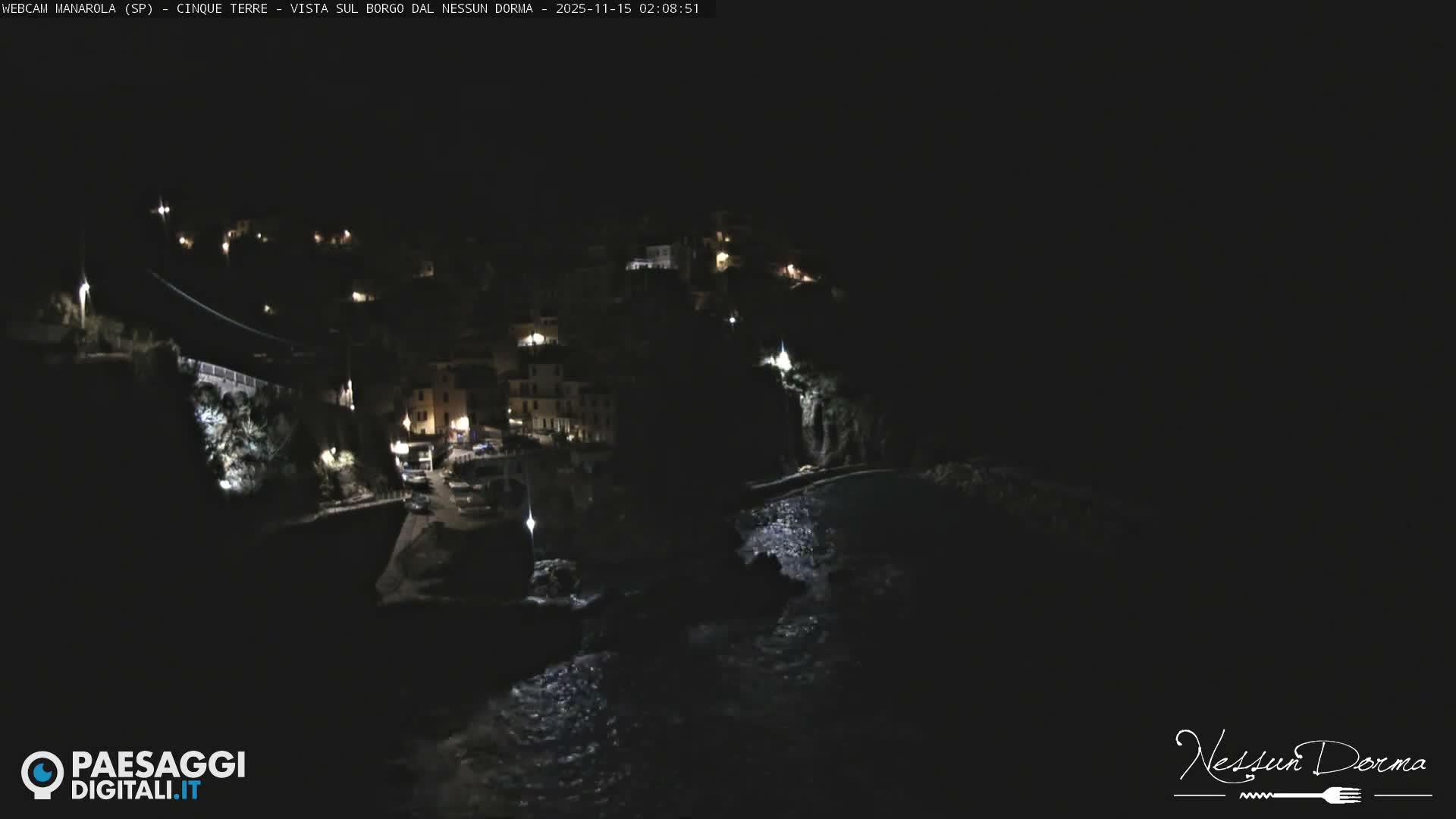 A clear night reveals a brightly lit coastal town nestled into a hillside overlooking a dark, calm sea.