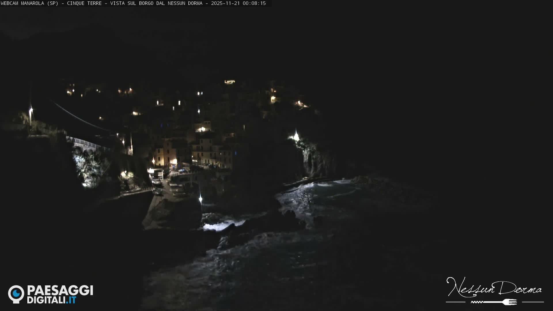 A clear night reveals a brightly lit coastal town nestled into a hillside overlooking a dark, calm sea.