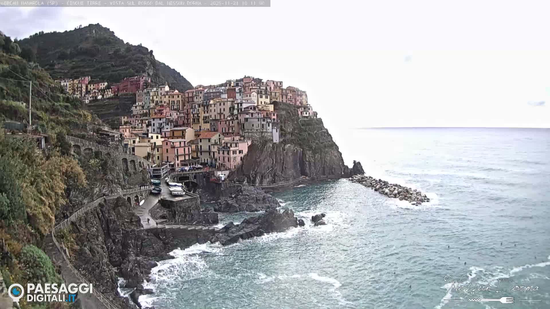 A picturesque coastal village with brightly colored buildings clinging to a steep, rocky hillside overlooks a choppy, greenish-blue sea under an overcast sky.