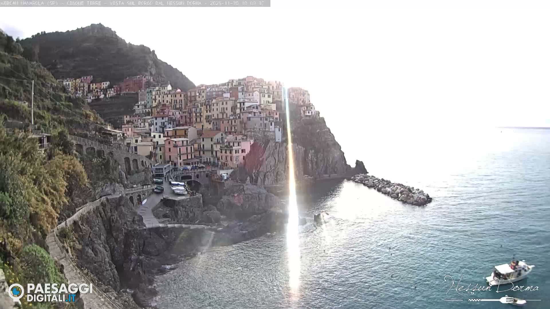 A vibrant, colorful town is built into steep cliffs overlooking a calm sea, intensely illuminated by a bright, sunny sky causing a prominent sun glare on the water.