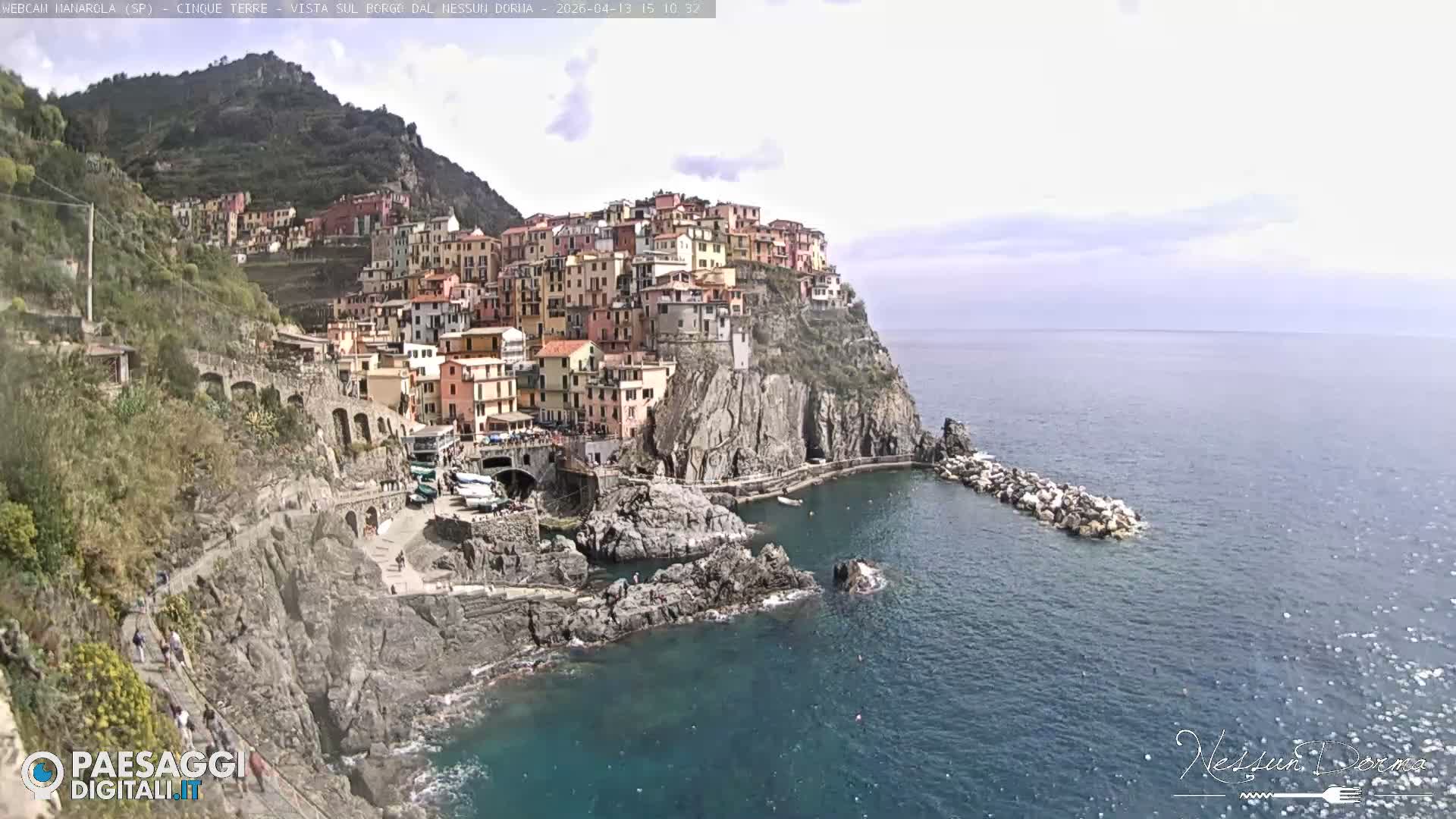A vibrant coastal village with colorful buildings is nestled into a steep, rocky hillside overlooking the calm, grey-blue sea under an overcast sky.