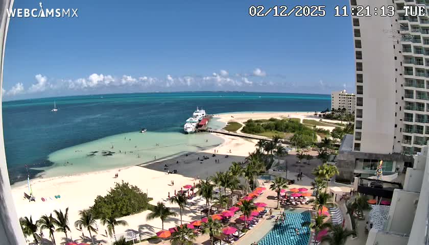 On a bright, sunny day with clear skies and scattered clouds, this image captures a bustling tropical beach with turquoise waters, a pier hosting ferries, people enjoying the sand and sea, a resort area with a pool and colorful umbrellas, all overlooked by a tall hotel building.