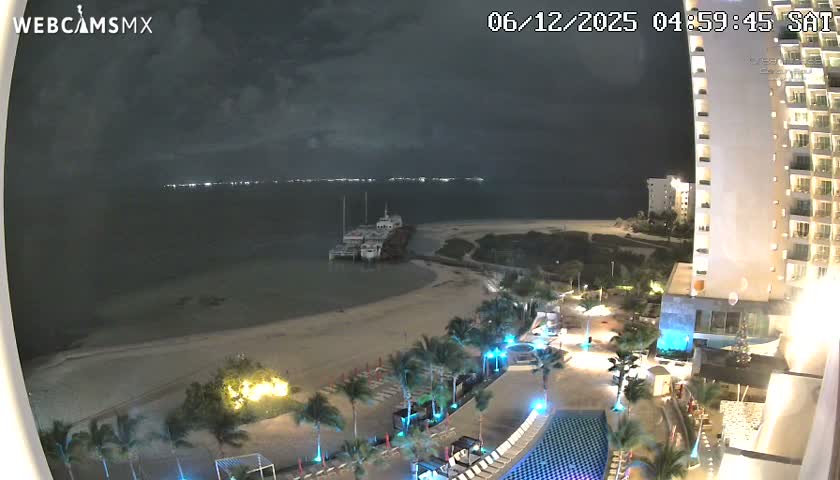 A pre-dawn view shows a brightly lit tropical resort beachfront with pools, palm trees, a sandy shore, and a pier with docked boats extending into the dark ocean, all under an overcast sky.