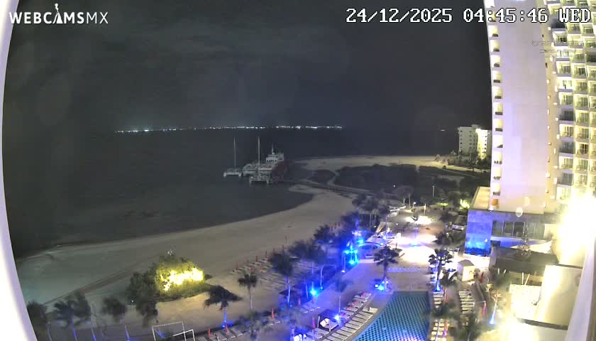A pre-dawn view shows a brightly lit tropical resort beachfront with pools, palm trees, a sandy shore, and a pier with docked boats extending into the dark ocean, all under an overcast sky.