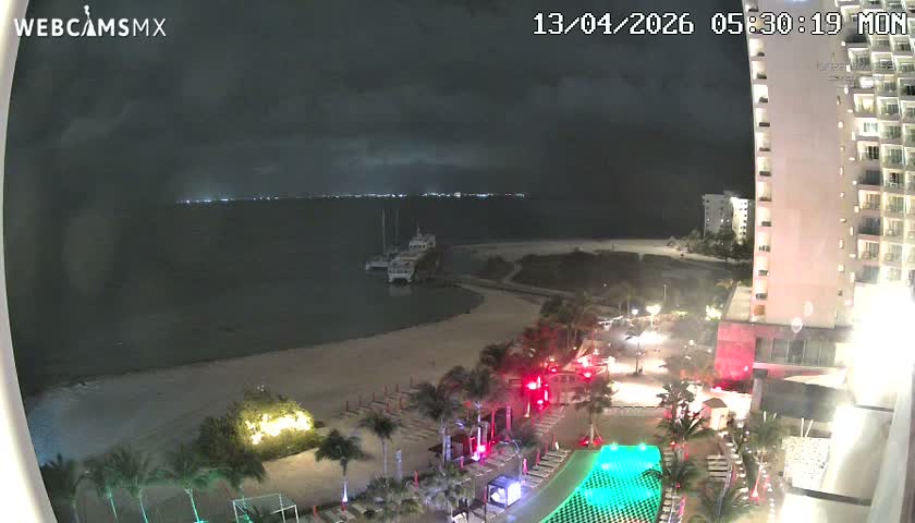 A pre-dawn view shows a brightly lit tropical resort beachfront with pools, palm trees, a sandy shore, and a pier with docked boats extending into the dark ocean, all under an overcast sky.