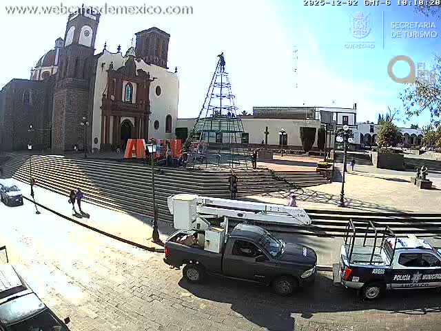 A sunny day illuminates a bustling town square featuring a historic church, people walking, a large metal structure being assembled, and two parked utility vehicles.