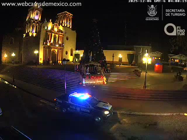 A police pickup truck with flashing blue and red lights is parked on a dark street at night, with a brightly illuminated historic church and steps in the background under clear weather conditions.