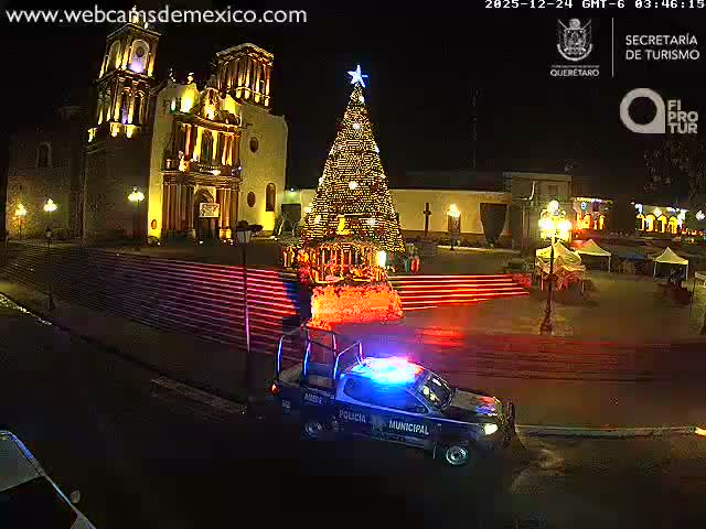 A police pickup truck with flashing blue and red lights is parked on a dark street at night, with a brightly illuminated historic church and steps in the background under clear weather conditions.