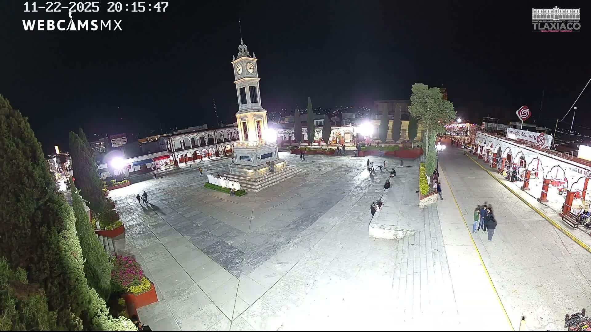 A brightly lit town square is seen at night, featuring a tall clock tower in the center, surrounded by arcaded buildings and trees, with several people strolling across the plaza under clear skies.