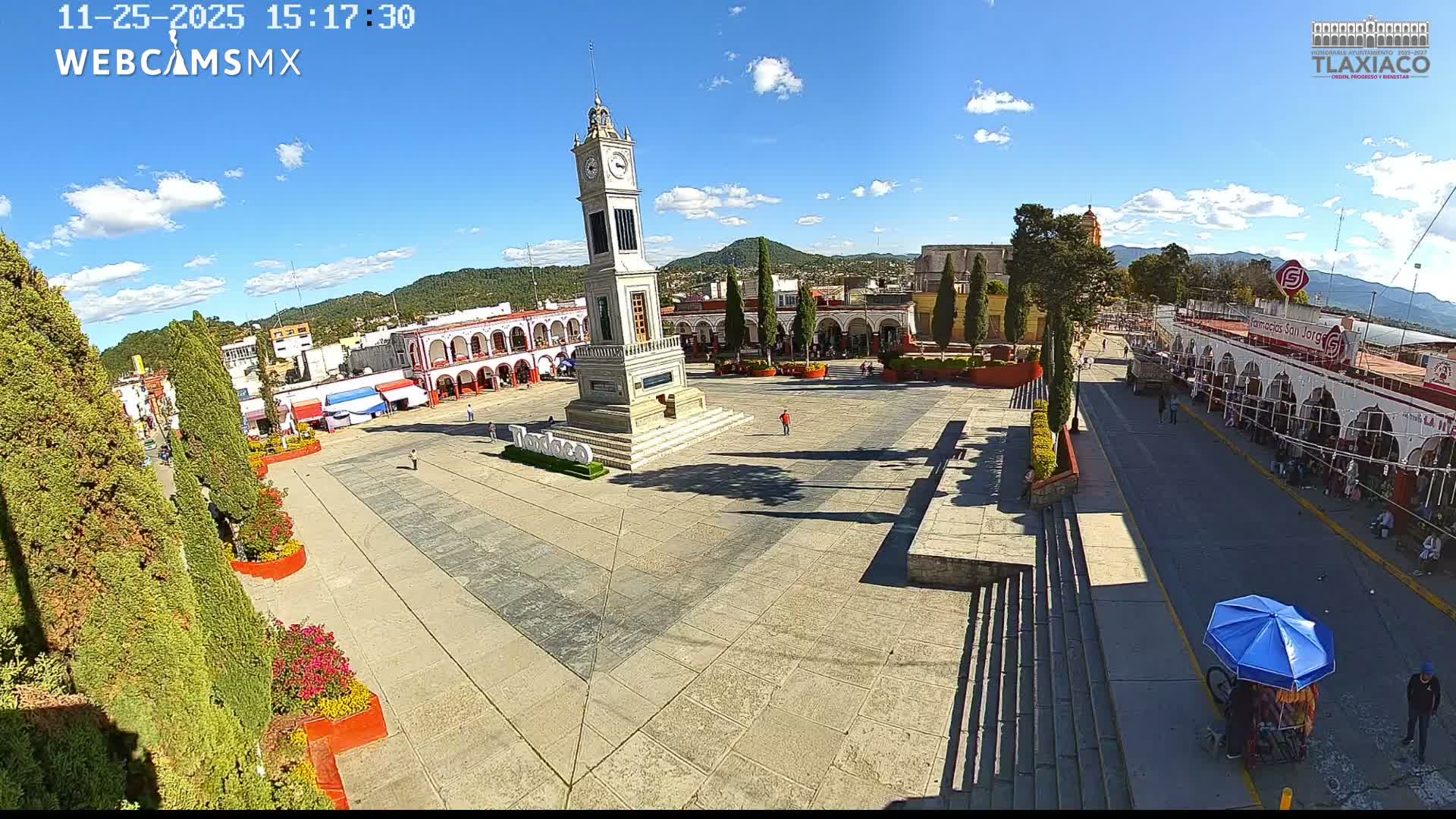 A brightly lit town square is seen at night, featuring a tall clock tower in the center, surrounded by arcaded buildings and trees, with several people strolling across the plaza under clear skies.