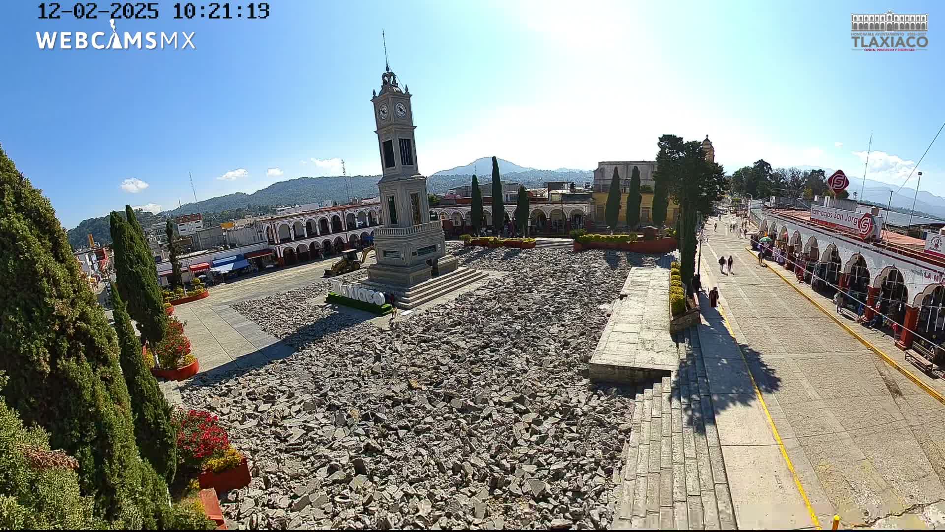 A sunny day with a clear blue sky reveals a large town square under apparent renovation, centered around an ornate clock tower and filled with broken pavement, bordered by arcaded buildings, a street with pedestrians, and distant green mountains.