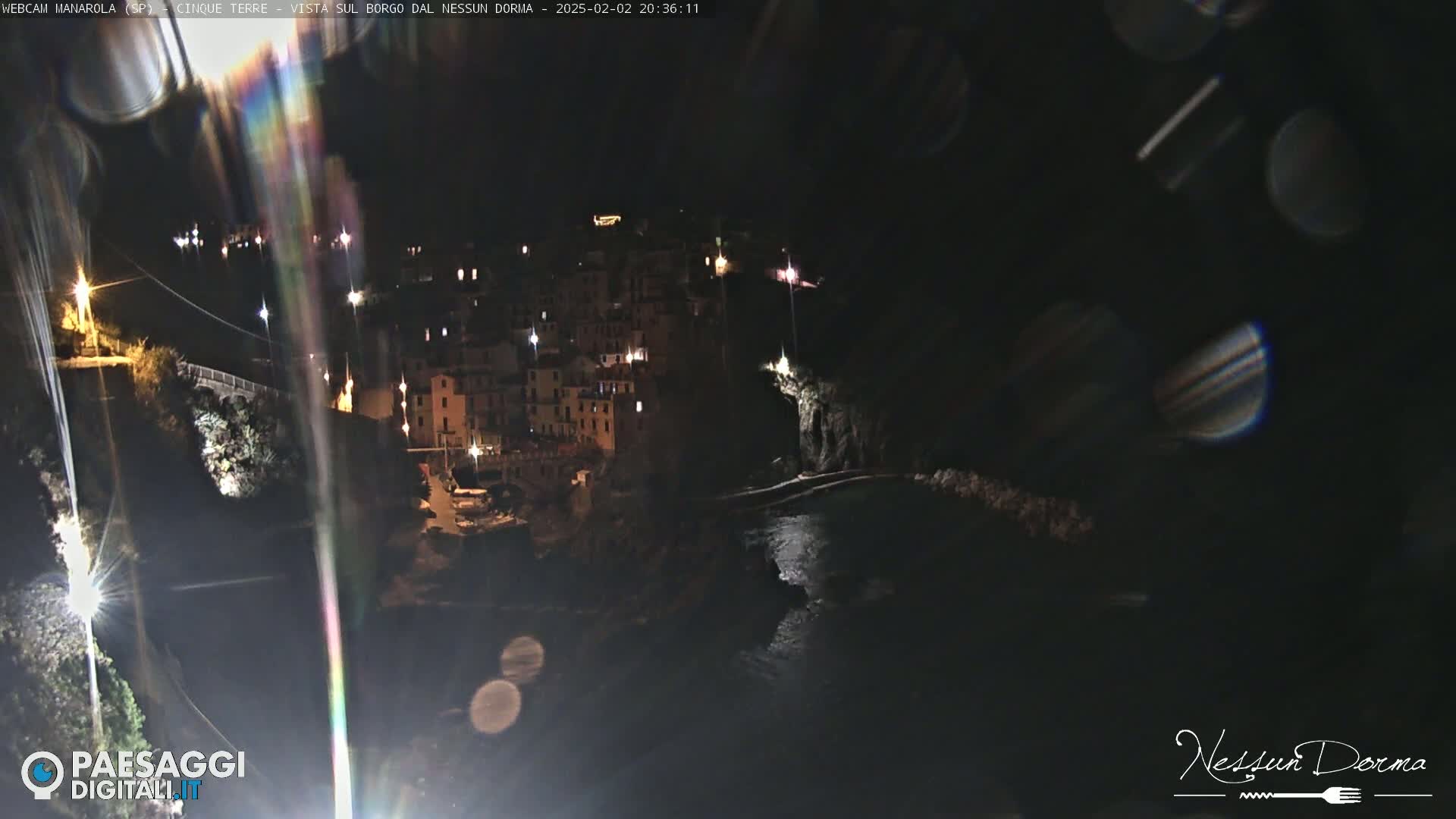 A nighttime, long-range view of a coastal town with buildings illuminated, situated on dark cliffs beside a body of water.