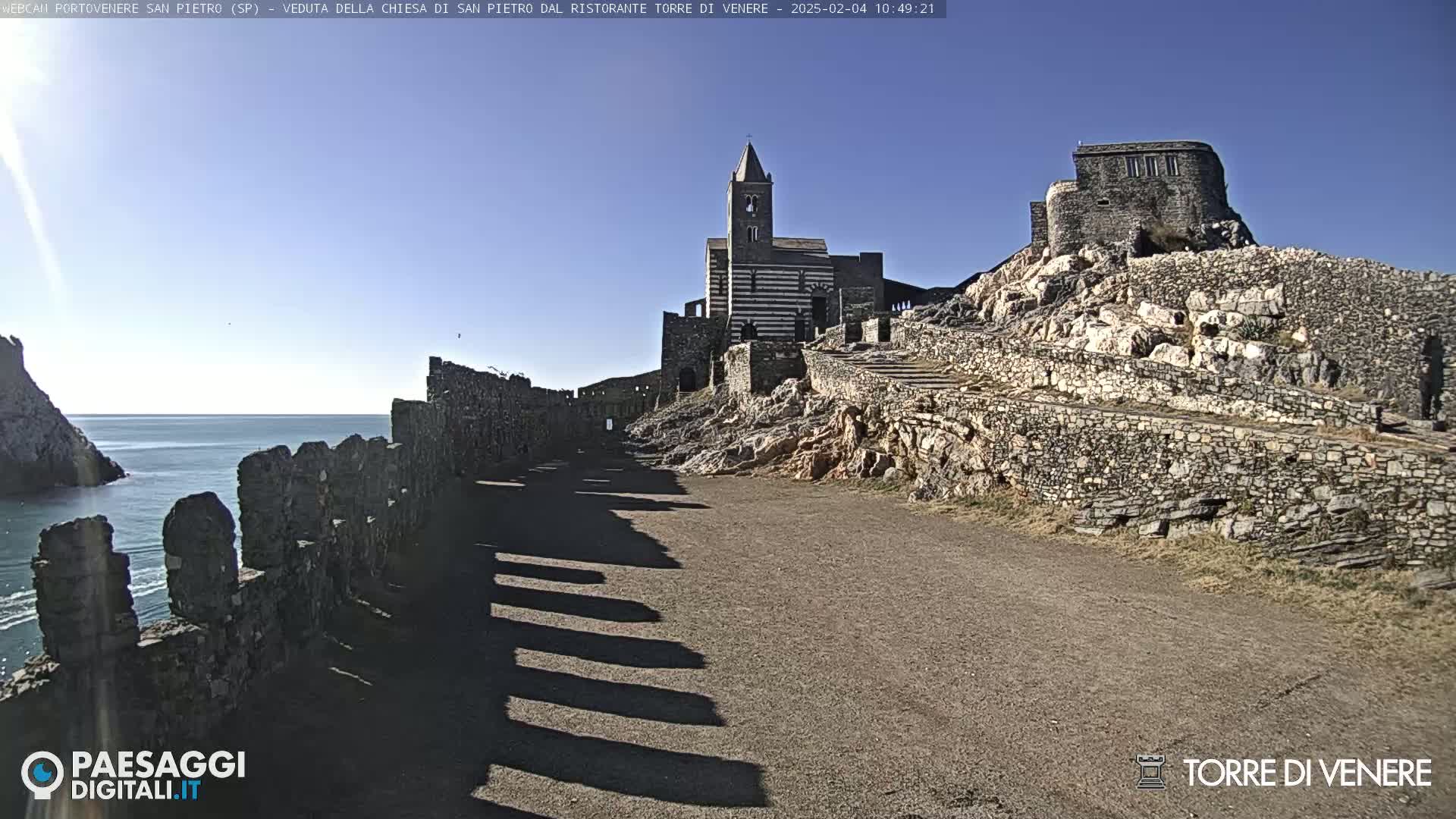 A stone church and castle ruins sit atop a rocky hill overlooking a calm sea under a clear blue sky.