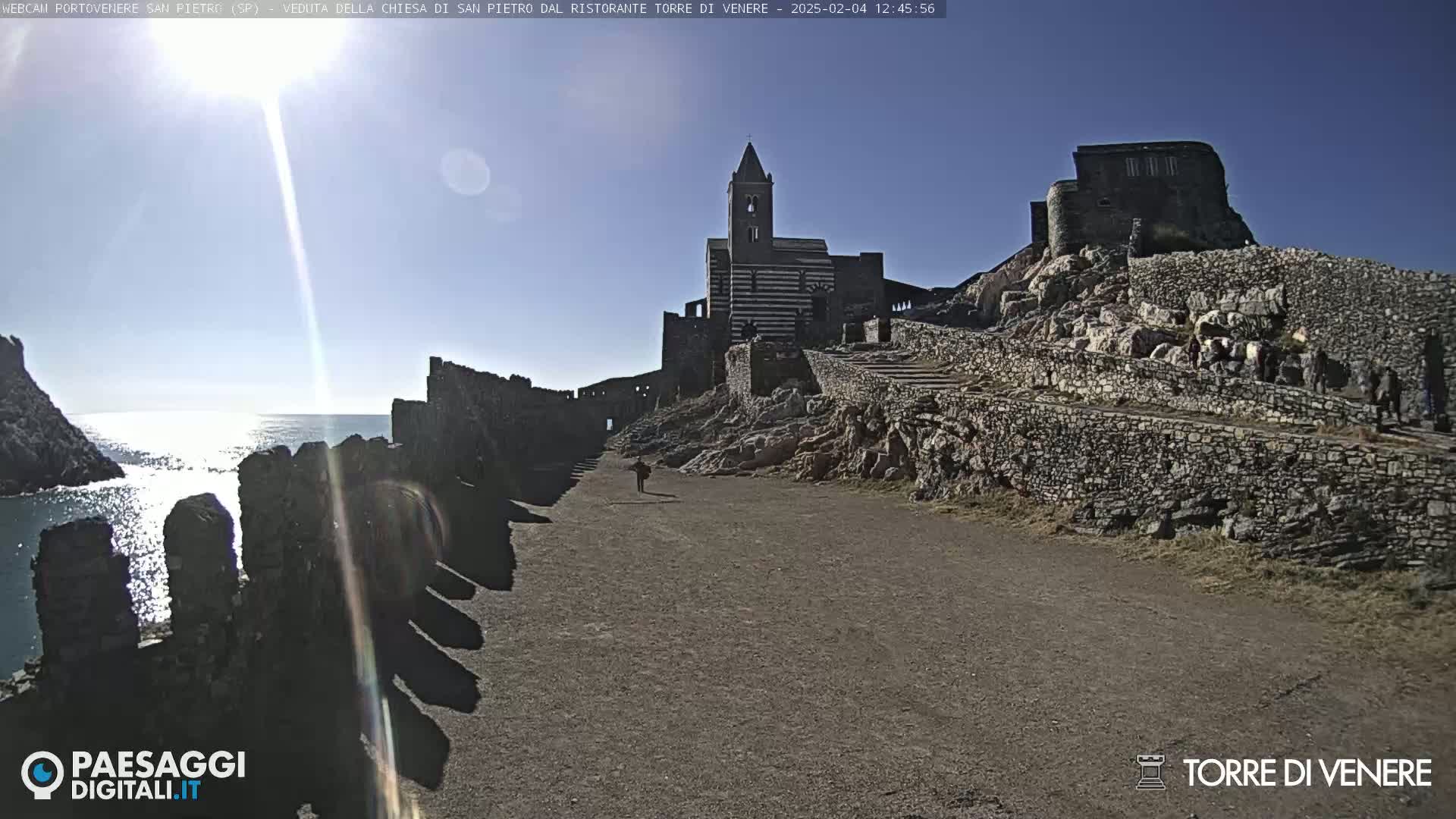 A stone path leads to a striped church and castle ruins perched atop a rocky cliff overlooking a calm sea under a sunny sky.