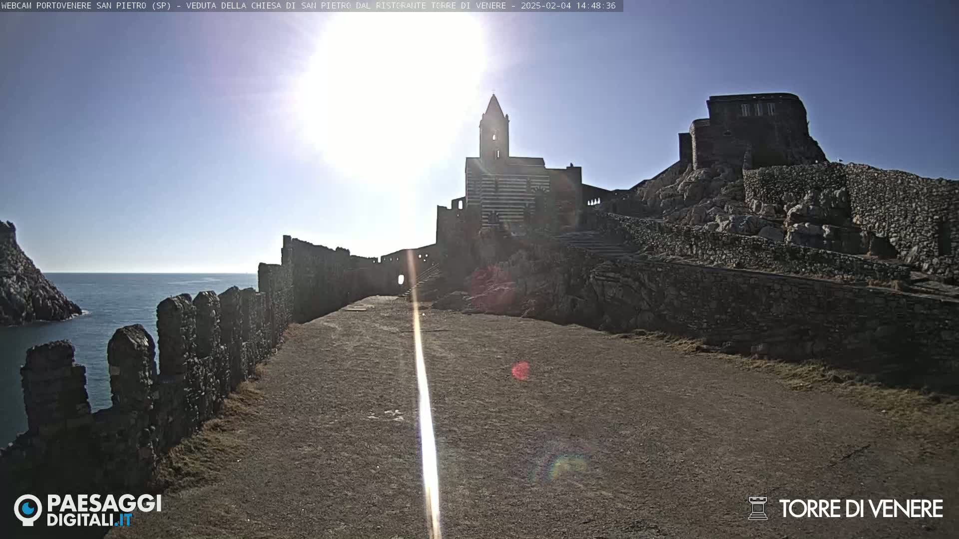 A stone pathway leads to a striped church and stone ruins overlooking a calm ocean under a bright, sunny sky.