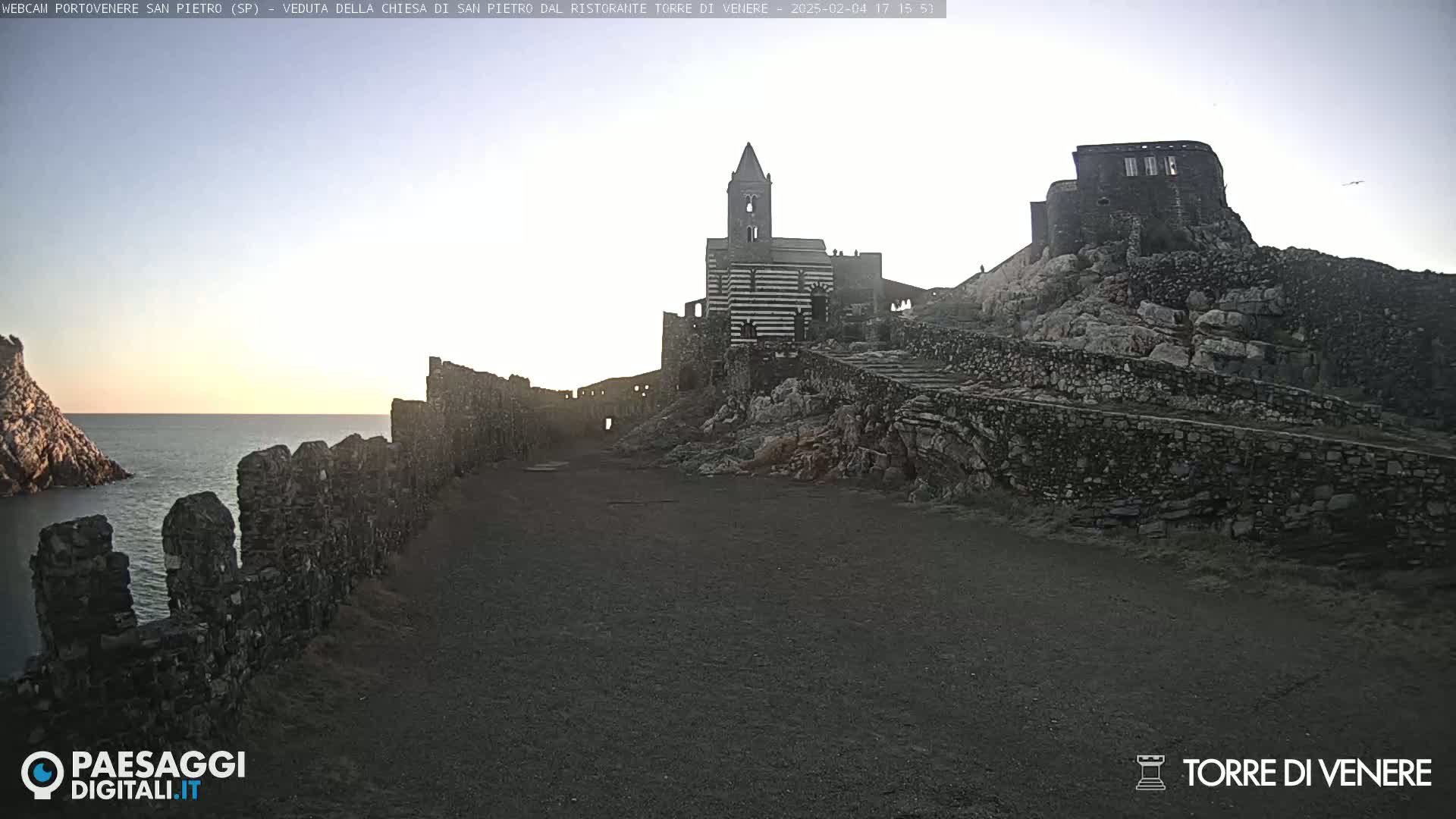 Stone ruins and a striped church stand on a rocky hillside overlooking a calm sea under a clear, light sky.
