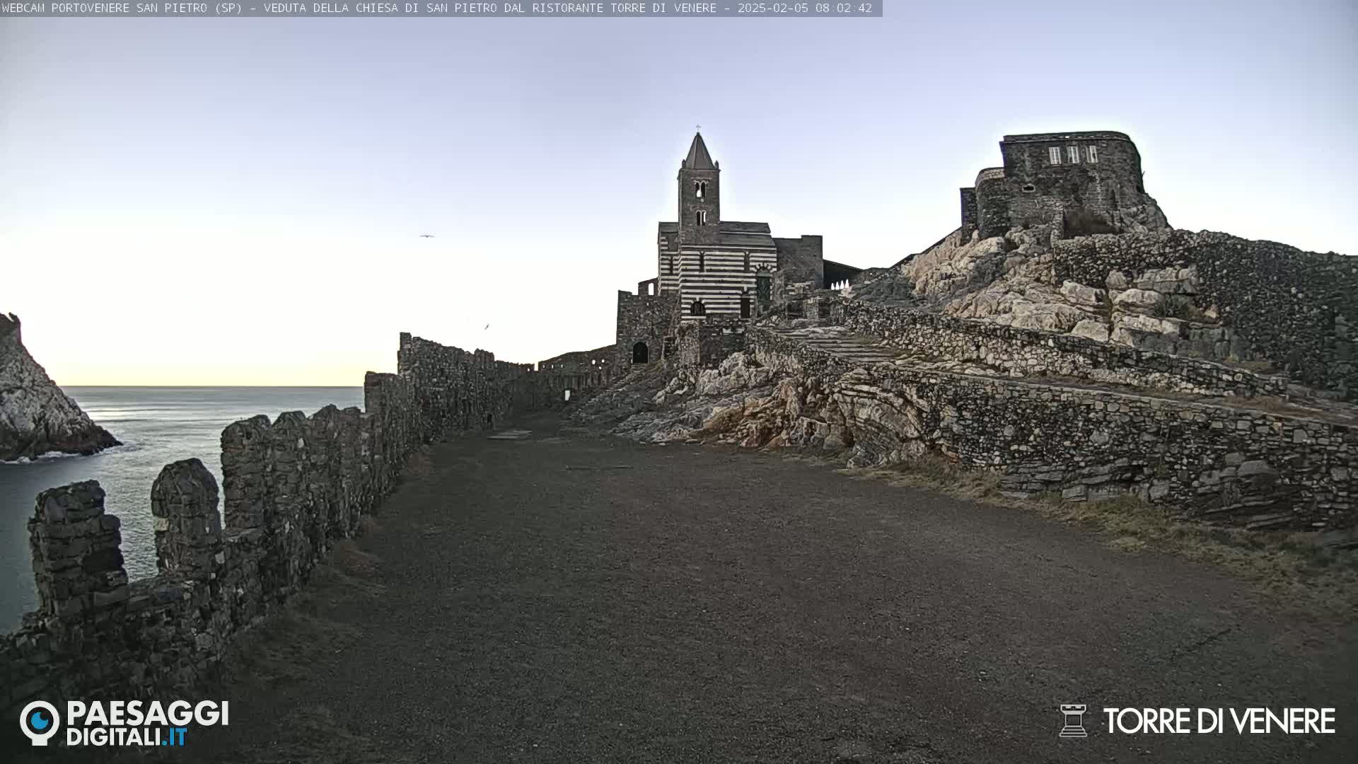 A stone church and fortress-like structures sit atop a rocky hill overlooking a calm sea under a clear, light-blue sky.