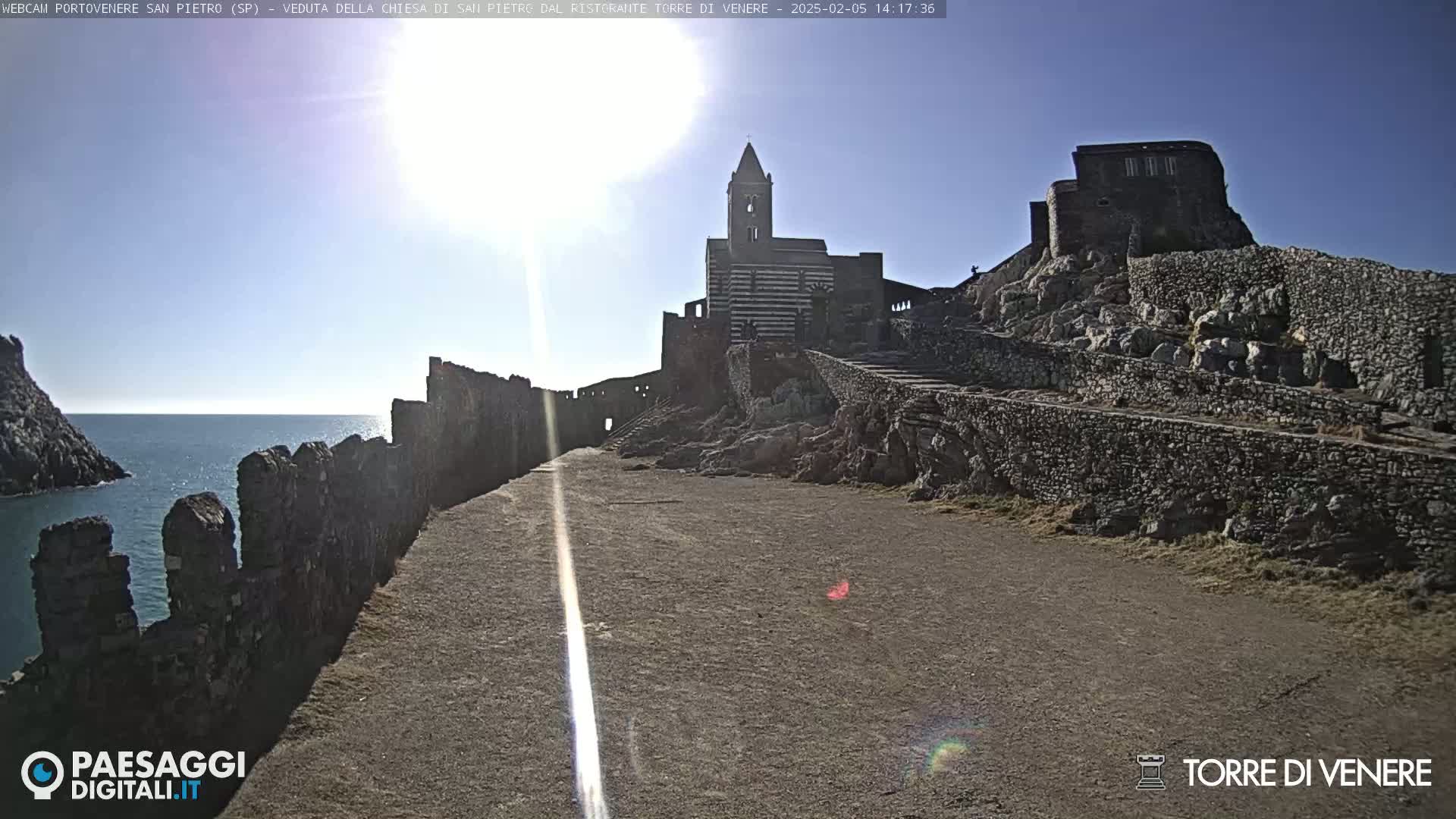 A stone path leads to a striped church and stone structures on a sunny day overlooking a calm sea.