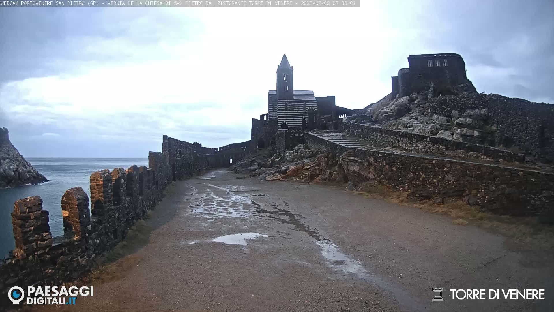A stone church and ruins are situated on a rocky hill overlooking a calm sea under a cloudy sky.