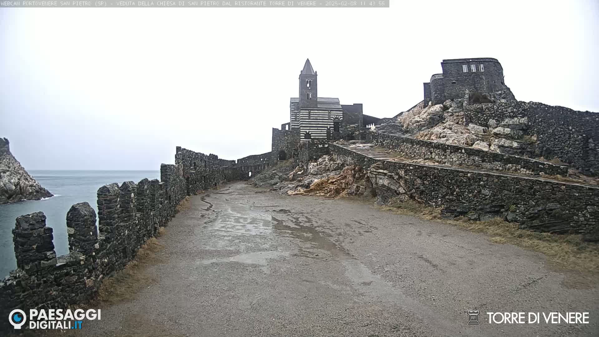 A wet, gray day reveals a stone pathway leading to a striped church and stone buildings atop a rocky hill overlooking a calm body of water.