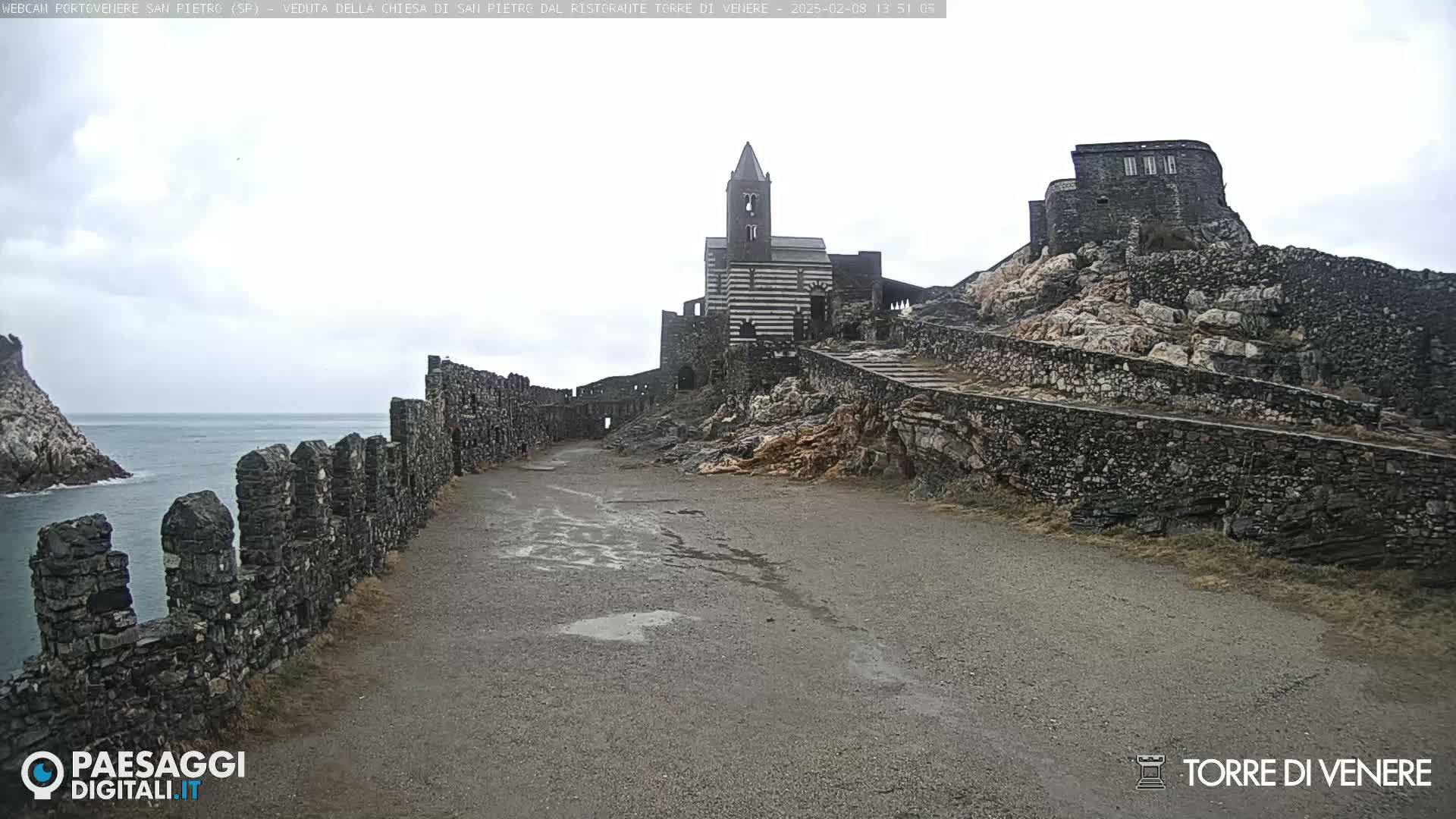 A stone path leads to a church and ruins situated on a rocky cliff overlooking a calm sea under an overcast sky.