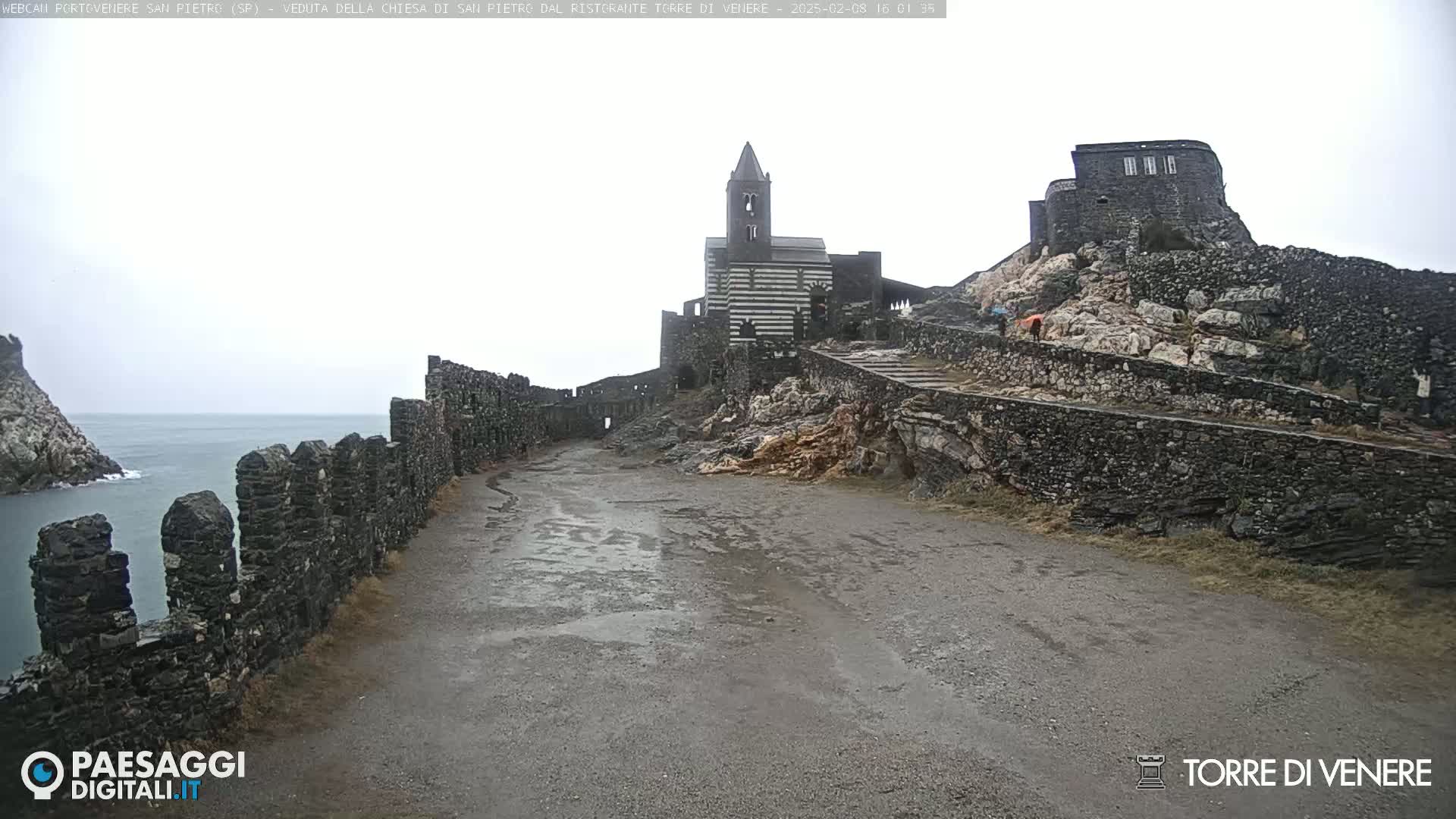 A wet, overcast day reveals a stone pathway leading to a striped church and castle ruins perched atop a rocky cliff overlooking the sea.