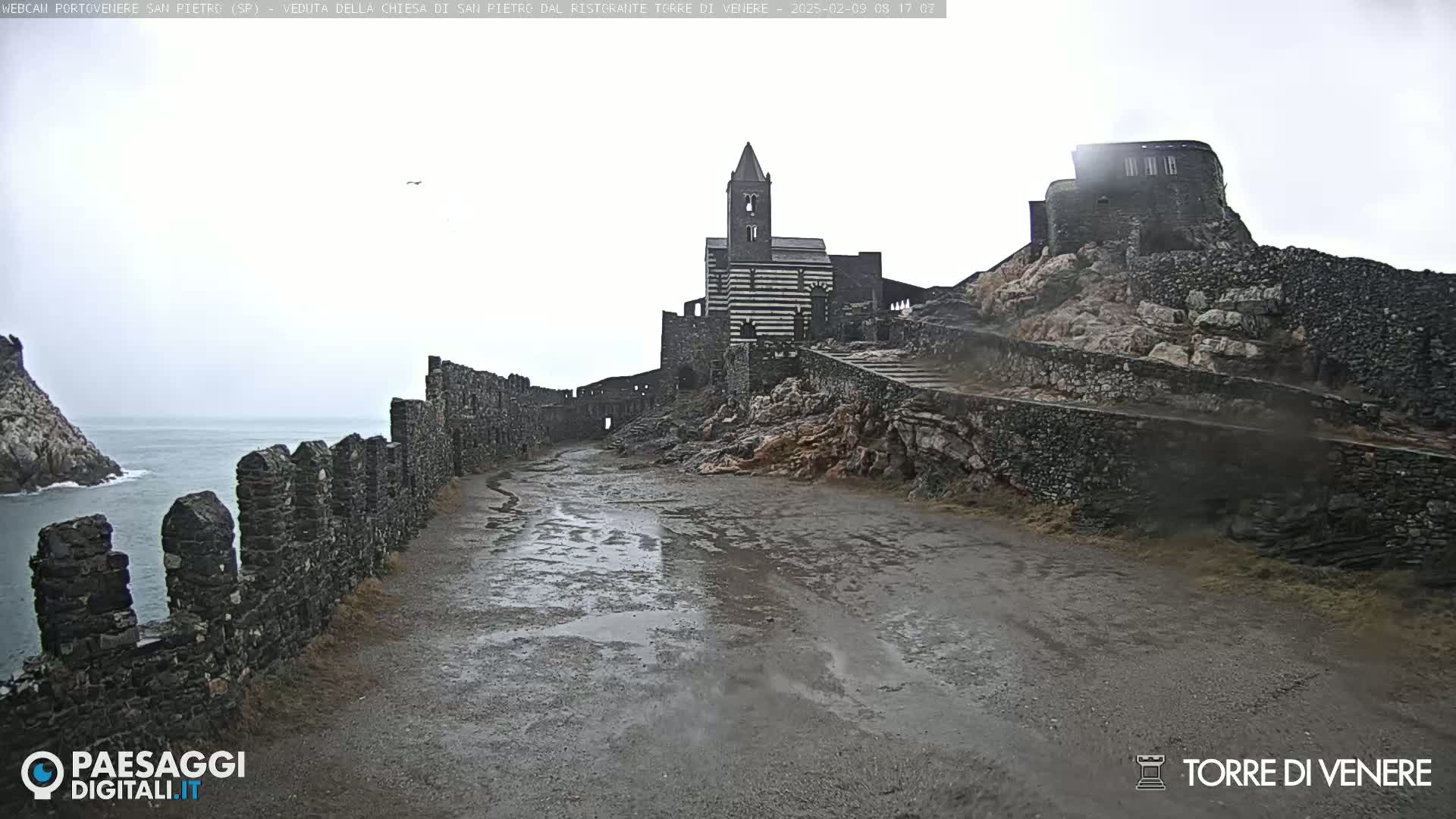 A wet, overcast day reveals a stone pathway leading to a striped church and ruins atop a rocky hill overlooking the ocean.