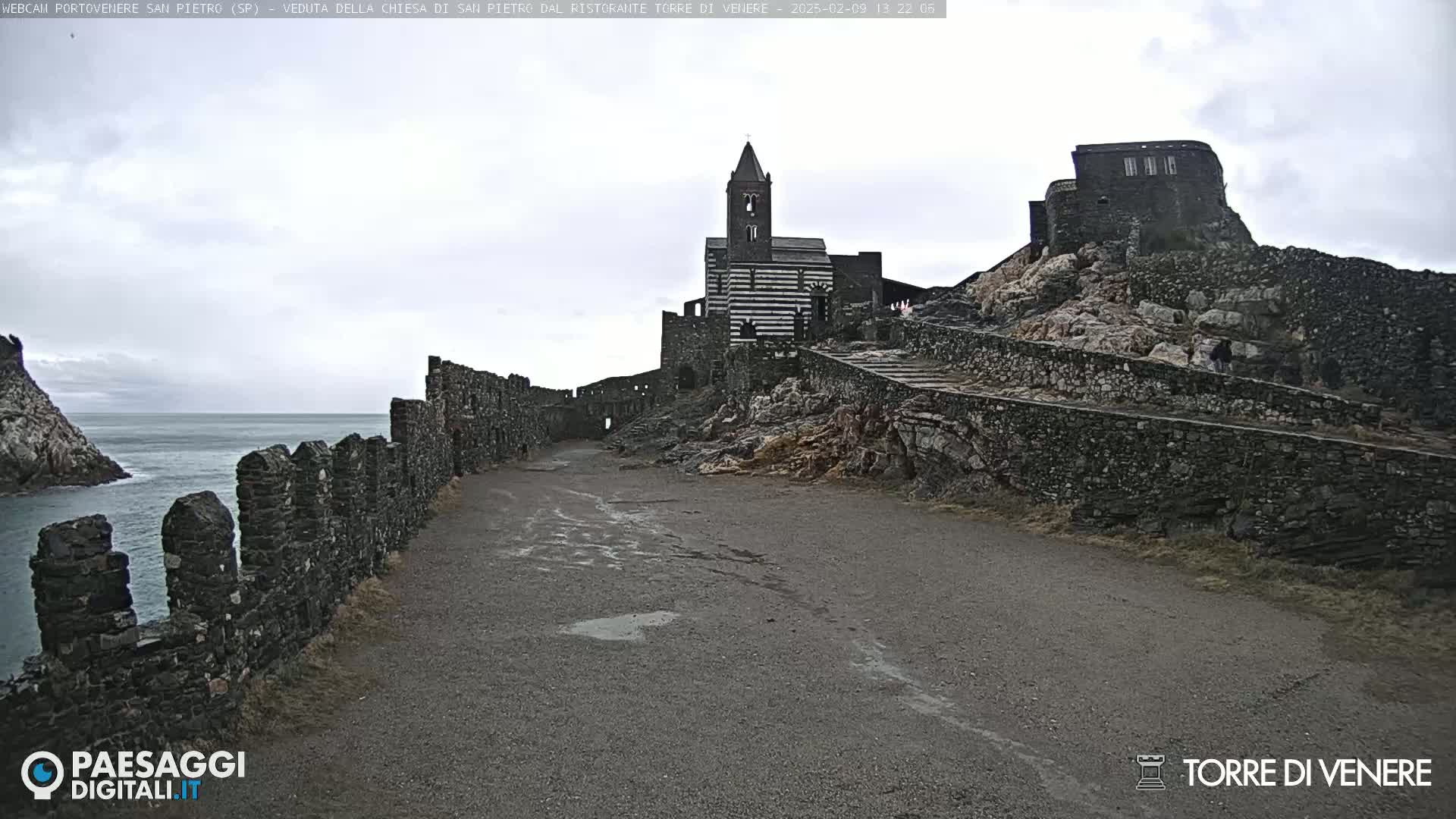 A stone path leads toward a striped stone church and castle ruins on a rocky hill overlooking a calm sea under an overcast sky.