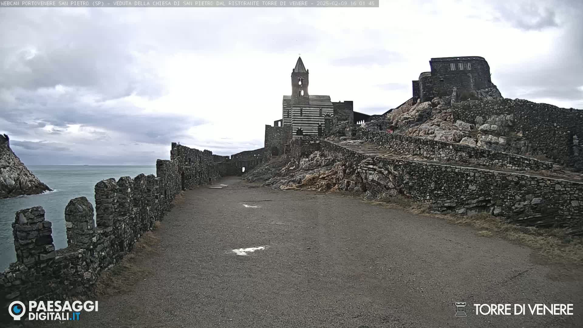 A stone path leads toward a striped church and castle ruins perched on a rocky cliff overlooking a calm sea under an overcast sky.