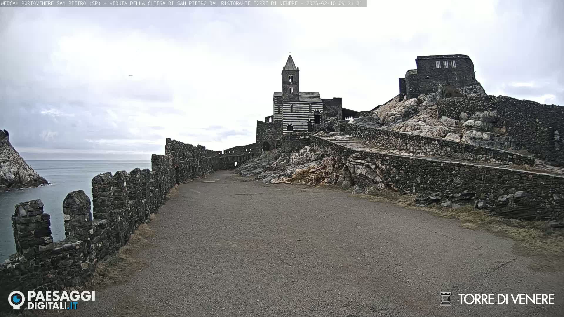 A stone path leads toward a striped church and stone buildings situated on a rocky cliff overlooking a calm sea under an overcast sky.