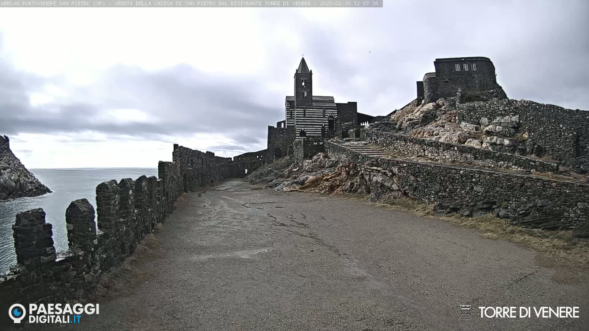 A stone path leads toward a striped church and castle ruins perched atop a rocky cliff overlooking a calm ocean under a cloudy sky.