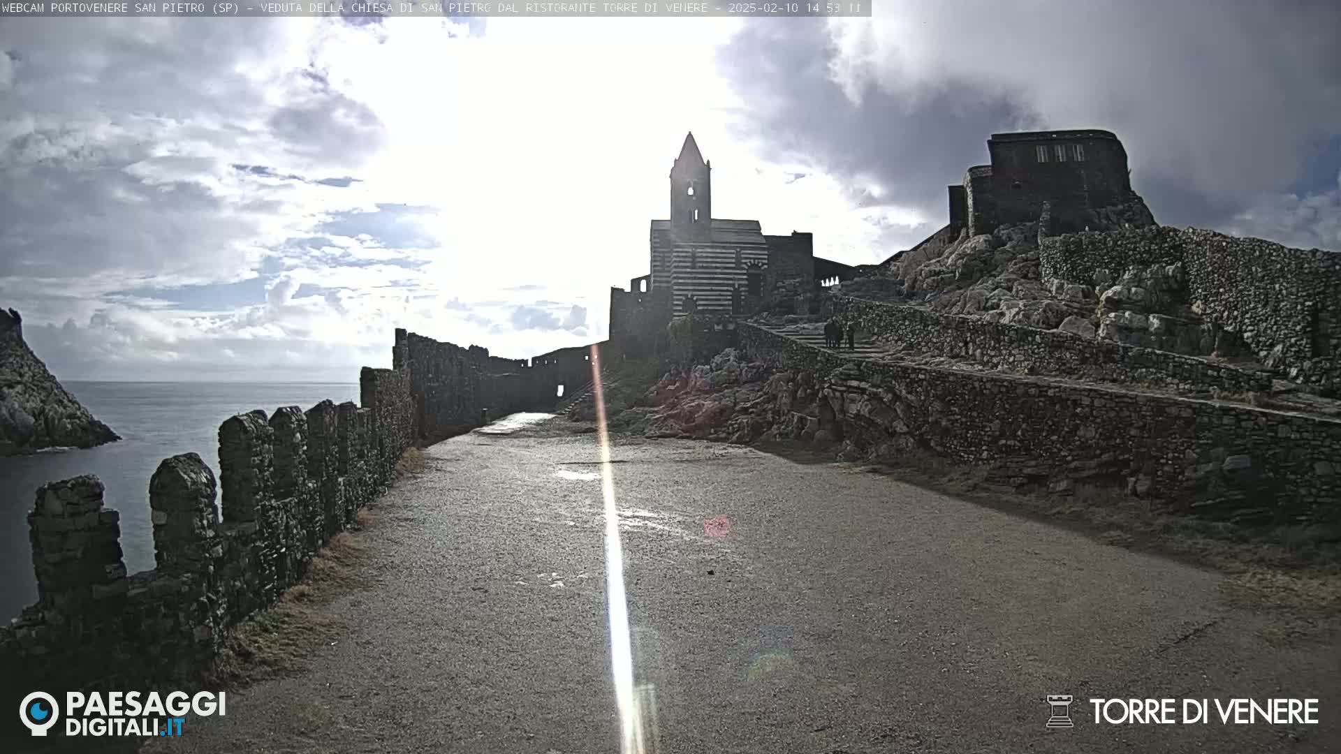 A stone path leads to a church and castle ruins on a rocky hillside overlooking a calm sea under a partly cloudy sky.