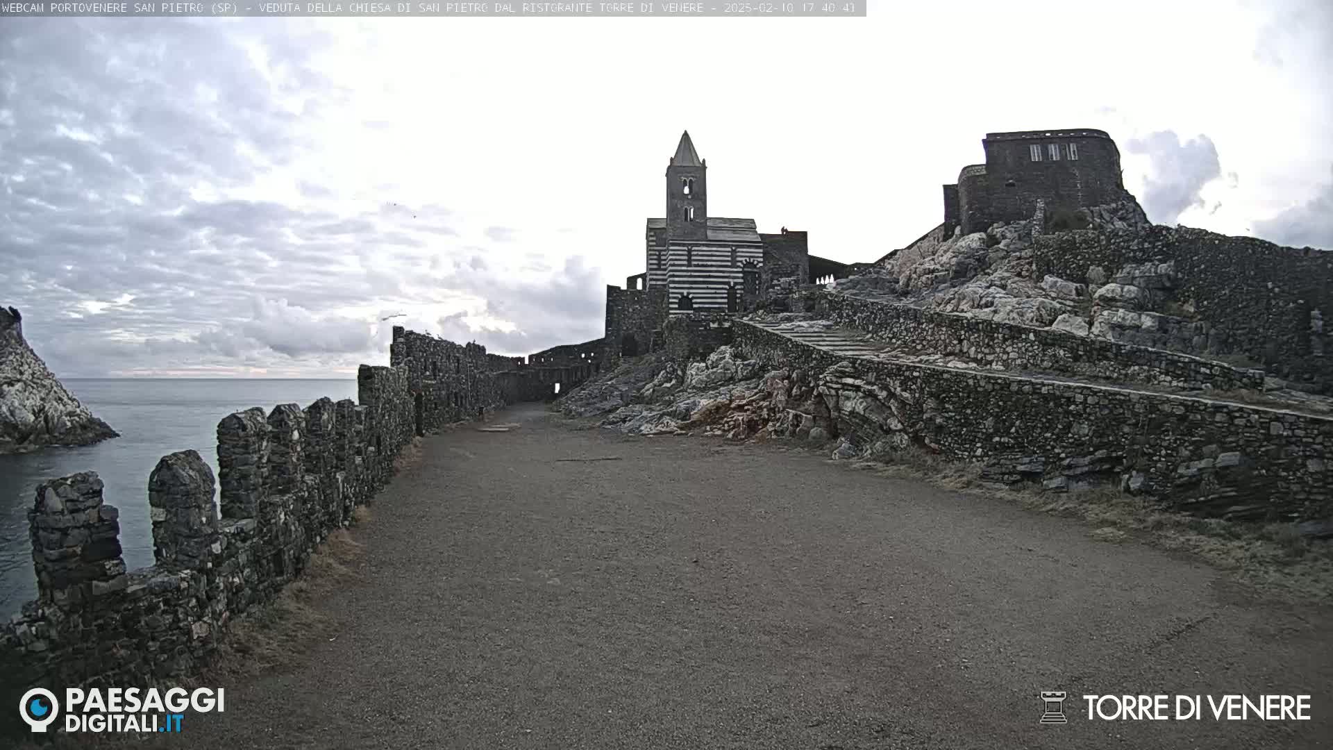 A stone path leads to a striped church and castle ruins on a rocky cliff overlooking a calm sea under a cloudy sky.