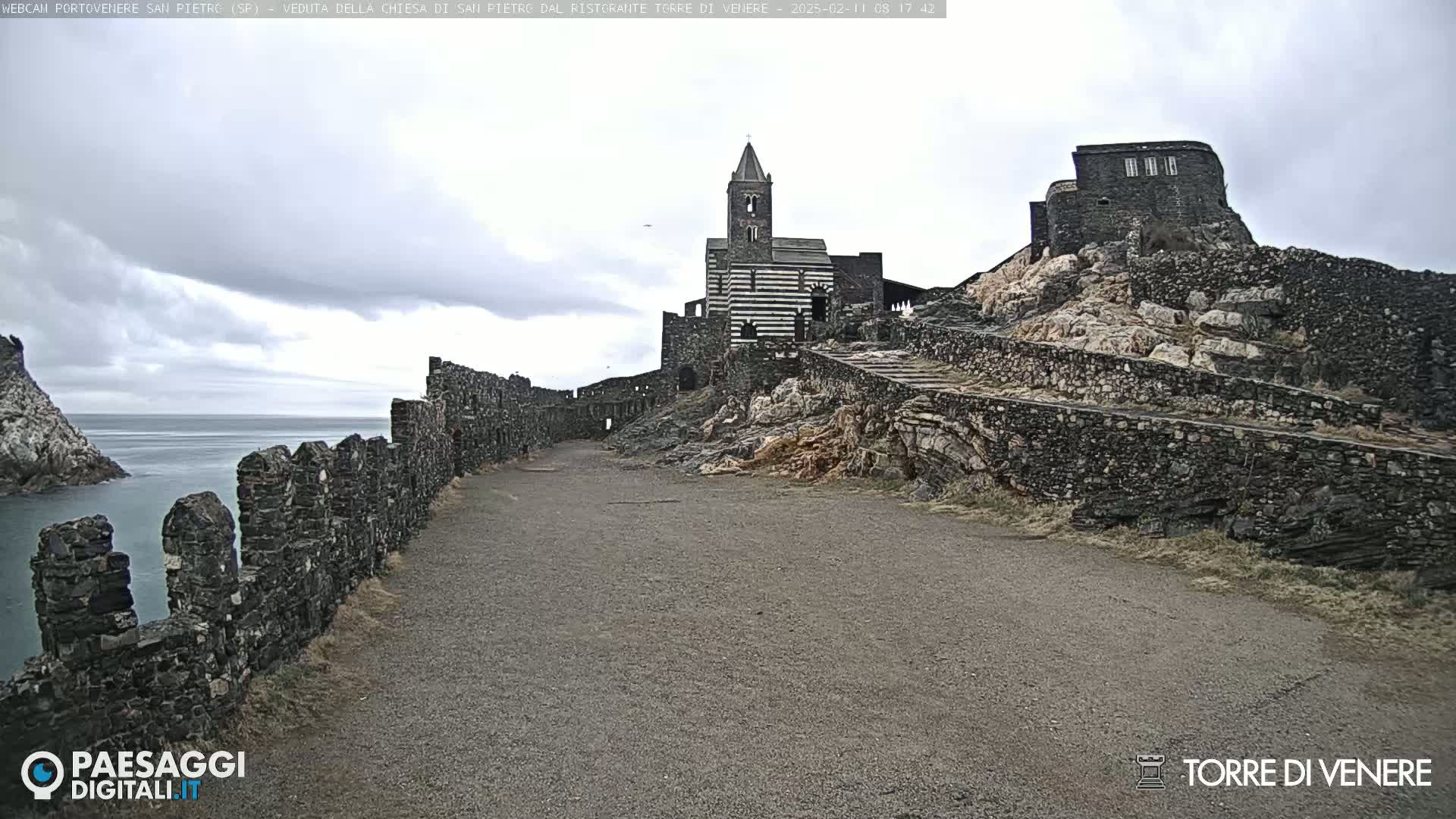 A stone church and fortress are situated on a rocky outcrop overlooking a calm sea under a cloudy sky.